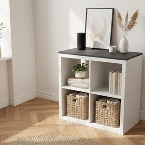 May include: A white and black cube storage unit with woven baskets, books, and decorative items. A black speaker sits on top. A framed abstract art print and a vase with dried plants are also displayed.