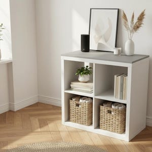 May include: A white cube storage unit with a gray top, holding decorative items. It features woven baskets, books, and plants. A black speaker, framed art, and a vase with dried flowers sit on top. The room has hardwood floors.
