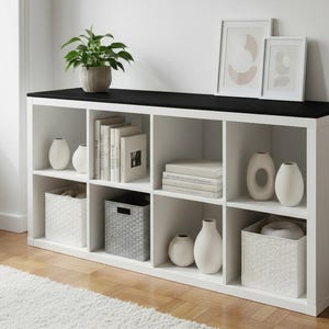 May include: A white cube storage unit with a black top, filled with books, vases, and decorative baskets. A potted plant and framed art pieces sit on top. The unit is on a hardwood floor.