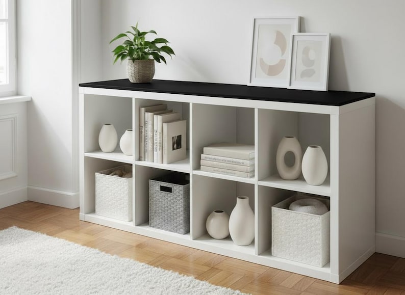 May include: A white cube storage unit with a black top, filled with books, vases, and decorative baskets. A potted plant and framed art pieces sit on top. The unit is on a hardwood floor.