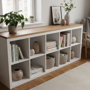 May include: A white storage unit with a light wood top, featuring multiple cubbies filled with books, vases, and decorative baskets. A potted plant and framed artwork sit on top. The unit is placed near a window and a neutral-toned rug.