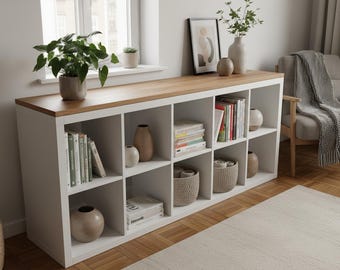 May include: A white storage unit with a light wood top, containing cubbies filled with books, vases, and baskets. A potted plant and framed artwork sit on top. The unit is near a window and a neutral rug. The unit is approximately 150 cm wide.