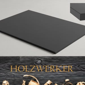 May include: A rectangular, matte black wooden board. The board is displayed on a light gray surface, with a close-up of the edge in the upper right corner. The word "HOLZWERKER" is displayed in gold lettering on a dark wood background.