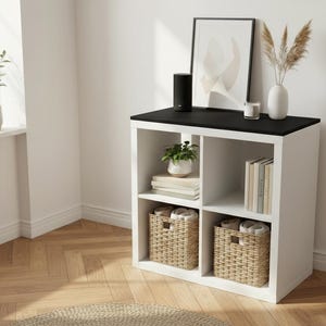 May include: A white cube storage unit with a black top, holding decorative items. The unit has two woven baskets, books, and a small potted plant. A black speaker, framed art, and a vase with dried plants sit on top.