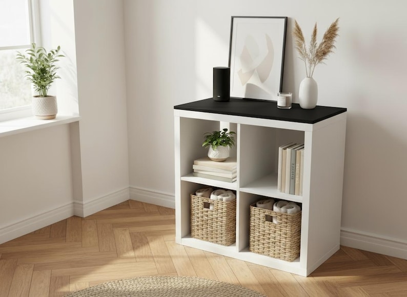 May include: A white cube storage unit with a black top, holding decorative items. The unit has two woven baskets, books, and a small potted plant. A black speaker, framed art, and a vase with dried plants sit on top.