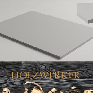 May include: A rectangular, light gray wooden panel with a smooth surface. The panel is displayed on a white background, with a smaller, angled section of the panel visible in the upper right corner. The word "HOLZWERKER" is displayed in gold lettering.