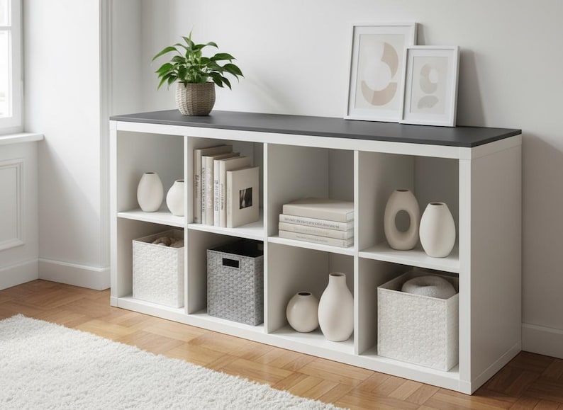 May include: A white cube storage unit with a dark gray top, filled with books, vases, and decorative baskets. A potted plant and framed art pieces sit on top. The unit is on a hardwood floor with a white rug.