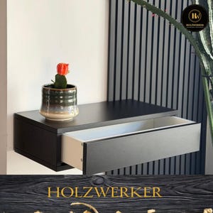 Könnte beinhalten: Ein schwarzer, schwebender Nachttisch mit einer offenen Schublade, die einen weißen Innenraum zeigt. Oben steht ein kleiner Topf mit einem Kaktus mit roter Blüte. Der Nachttisch ist an einer weißen Wand montiert, mit einer dunklen vertikalen Platte im Hintergrund.