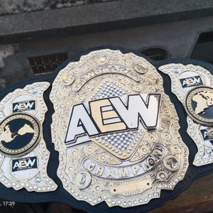 Könnte beinhalten: Ein gold-silberner AEW World Championship Gürtel. Die Mittelplatte zeigt die Buchstaben "AEW" in fetter Schrift. Der Gürtel hat Seitenplatten mit dem AEW-Logo und einer Weltkarte.