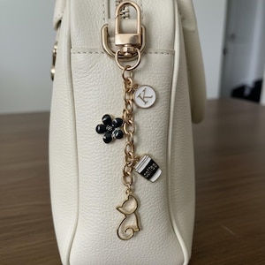 May include: A cream-colored handbag with a gold-tone zipper and hardware. A gold chain charm hangs from the zipper, featuring a black flower, a coffee cup, a cat silhouette, and a white circle with the letter "K".