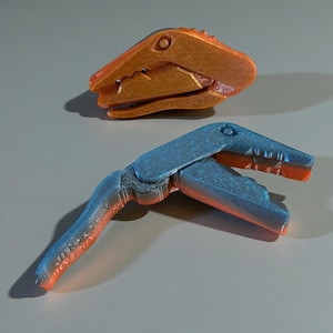 Op de afbeelding: Twee 3D-geprinte dinosaurustools. Eén is een dinosaurushoofd, de andere is een blauw en oranje klauwachtig gereedschap. De klauw is open en onthult het tandachtige ontwerp. De tools liggen op een grijze ondergrond.
