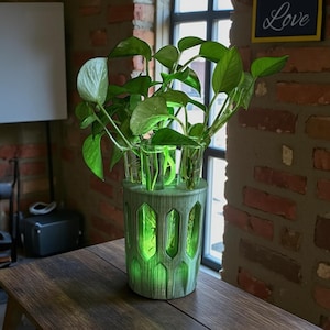 Op de afbeelding: Een groene decoratieve vaas met een uniek ontwerp, met daarin verschillende kleine glazen vazen gevuld met groene bladplanten. De vaas staat op een houten tafel, met een bakstenen muur en een raam op de achtergrond.