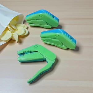 May include: Three green and blue alligator-shaped bag clips are displayed, with a bag of potato chips in the background. The clips are made of plastic and have a hinged design for sealing bags. The alligator clips are a fun and functional kitchen accessory.