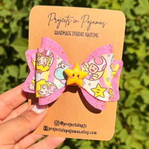 May include: A handmade pink glitter hair bow with a yellow star centre and a colourful cartoon print. The bow is attached to a brown card with the text "Projects in Pajamas" and "Handmade During Naptime".