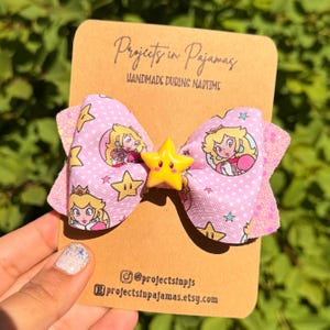 May include: A pink bow featuring Princess Peach and star details, with a yellow star centre. The bow has a polka dot pattern. The card reads "Projects in Pajamas" and "Handmade During Naptime."