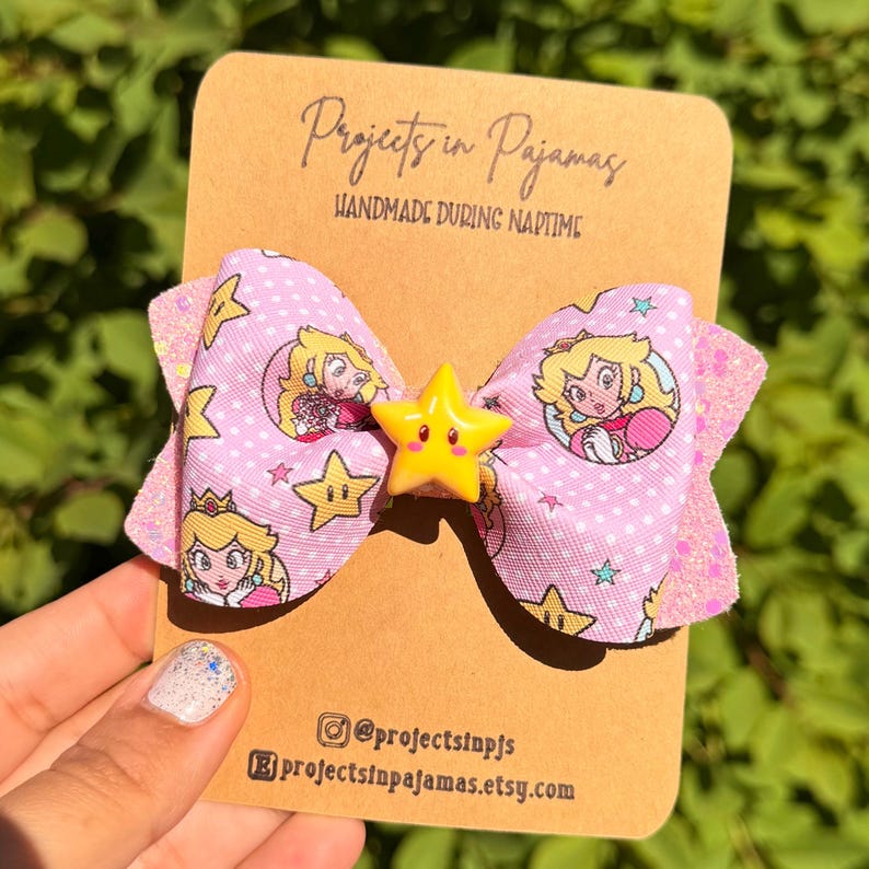 May include: A pink bow featuring Princess Peach and star details, with a yellow star centre. The bow has a polka dot pattern. The card reads "Projects in Pajamas" and "Handmade During Naptime."
