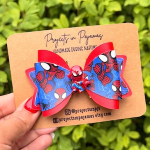 Könnte beinhalten: Eine Haarschleife mit blau-rotem Spider-Man-Muster und einem zentralen Spider-Man-Anhänger. Die Schleife ist auf einer beigen Karte befestigt, die mit dem Text Projects in Pajamas, HANDMADE DURING NAPTIME und Social-Media-Infos bedruckt ist.