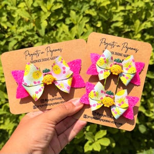 May include: Three hair bows on brown cardboard display cards labeled Projects in Pajamas. Each bow features a white faux leather layer with a yellow pineapple and pink bow pattern, accented by bright pink glitter fabric and a pineapple charm.