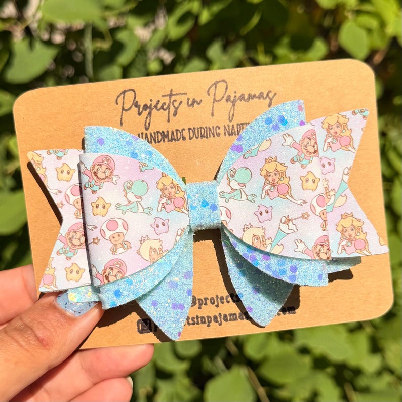 Puede incluir: Un lazo para el pelo hecho a mano con una base de purpurina azul claro y capas con un estampado de personajes de dibujos animados. El lazo est&aacute; sujeto a una tarjeta marr&oacute;n con el texto "Projects in Pajamas" y "handmade during naptime".