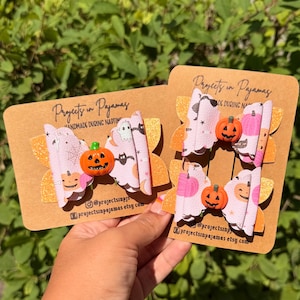 May include: Three hair bows attached to brown cardboard display cards labelled Projects in Pajamas. The bows feature a light pink pattern of ghosts, bats, and pumpkins with orange glitter backing and a small orange jack-o'-lantern centre accent.
