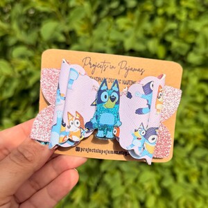 May include: A handmade hair bow featuring a cartoon character. The bow has a light pink background with a repeating pattern of cartoon characters. The center of the bow has a glittery blue cartoon character. The bow is attached to a brown card with text.