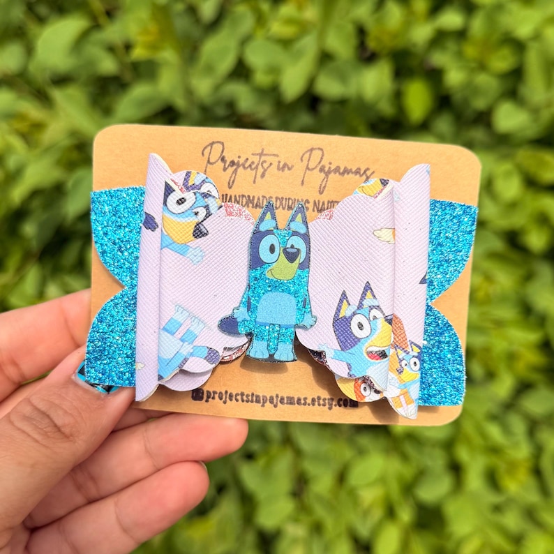 May include: A handmade hair bow featuring a cartoon character design. The bow has a light purple center with character graphics and bright blue glitter accents. The bow is attached to a brown card with text.