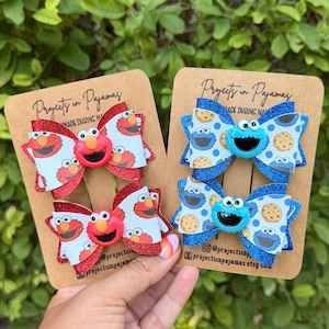 Peut inclure: Deux paires de nœuds pour cheveux fixés sur des cartes de présentation en carton brun marquées Projects in Pajamas. La paire de gauche présente des bords à paillettes rouges et un motif blanc de visages d'Elmo avec des centres Elmo rouges. La paire de droite présente des bords à paillettes bleues et un motif blanc de visages de Cookie Monster et de biscuits avec des centres Cookie Monster bleus.