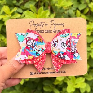 K&ouml;nnte beinhalten: Eine mehrlagige Haarschleife mit einem bunten Aufdruck von Mario- und Toad-Figuren, Regenb&ouml;gen und goldenen Sternen. Die Schleife ist mit leuchtend roten Glitzerstoffschichten verziert und auf einer beigen Pr&auml;sentationskarte von Projects in Pajamas befestigt.