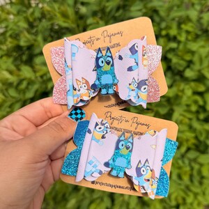 May include: Two handmade hair bows featuring a cartoon dog character in shades of blue, on a light purple background. The bows have glitter accents and are attached to brown cardstock with the text "Projects in Pajamas".
