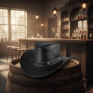 Könnte beinhalten: Ein schwarzer Leder-Cowboyhut mit geflochtenem Band und messingfarbenen Nieten liegt auf einem Holzfass. Der Hut hat eine breite Krempe, in einer schwach beleuchteten Bar.