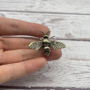 May include: A bronze-colored bee-shaped pin. The pin has detailed wings and body segments, with a textured surface. The bee is a decorative accessory, suitable for clothing or accessories.