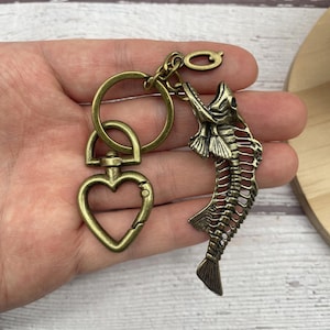 May include: Antique brass-colored keychain featuring a fish skeleton charm. The keychain includes a heart-shaped clasp, a large ring, and a small ring. The fish skeleton is detailed with visible ribs and fins.