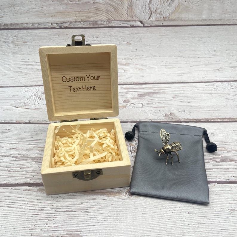 May include: A small, open wooden box with the text "Custom Your Text Here" engraved on the inside lid. The box contains shredded paper. A bronze-colored bee-shaped pin rests on a gray drawstring bag.