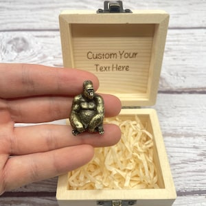Könnte beinhalten: Eine kleine, bronzefarbene Gorillafigur sitzt in einer offenen Holzkiste, die mit geschreddertem Papier gefüllt ist. Die Kiste hat einen Klappdeckel und der Text "Custom Your Text Here" ist auf der Innenseite des Deckels eingraviert.