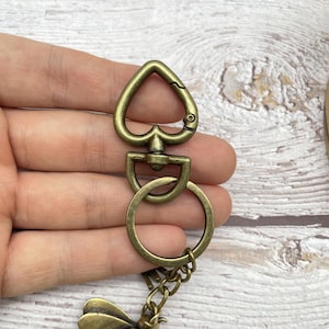 Pu&ograve; includere: Portachiavi color ottone anticato con chiusura a forma di cuore, anello a D e catenella. Il portachiavi &egrave; attaccato a un ciondolo a forma di ape. L'hardware ha un aspetto vintage e usurato.
