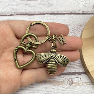 May include: Antique brass-colored keychain featuring a bee charm with detailed wings and a letter "W" pendant. Includes a heart-shaped clasp and a round keyring. The bee charm is approximately 1.5 inches long.