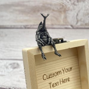 May include: A dark metal sculpture of a beetle-like creature sits on a small, light-coloured wooden box. The box has a hinged lid and the words "Custom Your Text Here" are engraved on the front. The creature has a large horn and detailed legs.