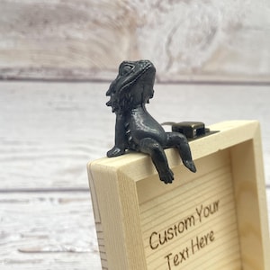 May include: A small, black lizard figurine perched on the edge of a light-colored wooden box. The box has a metal clasp and the words "Custom Your Text Here" engraved on the front. The lizard is detailed and appears to be looking up.
