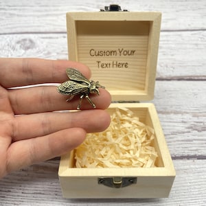 Może przedstawiać: Małe, otwarte drewniane pudełko z mosiężną figurką pszczoły. Pudełko jest jasne z napisem "Custom Your Text Here" w środku. Pszczoła jest szczegółowa ze skomplikowanymi wzorami skrzydeł.