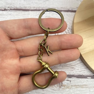 Op de afbeelding: Antieke messing sleutelhanger met een cirkelvormige ring, ketting en een 'K' initiaal bedel. De sleutelhanger heeft een decoratieve sluiting in de vorm van een beugel. Het ontwerp is vintage.