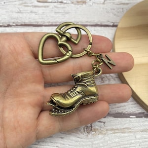 Puede incluir: Un llavero de color bronce con un dije en miniatura de bota de senderismo. El llavero incluye un cierre en forma de corazón y un dije con la letra "K". El dije de la bota tiene detalles intrincados, de aspecto vintage.
