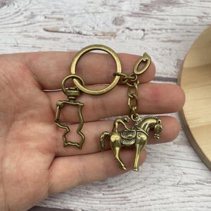 Puede incluir: Llavero de color latón antiguo con un caballo con jinete, un llavero y un dije decorativo. El caballo tiene una silla de montar y una brida. El llavero se sostiene en una mano.