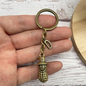 Könnte beinhalten: Ein bronzefarbener Erdnuss-Schlüsselanhänger mit einem lächelnden Gesicht und einem kleinen Hut. Der Schlüsselanhänger hat einen Ring und eine Kette mit einem kleinen Buchstaben "D" Anhänger. Die Erdnuss ist strukturiert und detailliert.