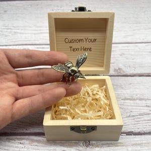 May include: A small, open wooden box with a brass-colored bee pin resting on a hand. The box is filled with shredded paper and has the text "Custom Your Text Here" engraved on the inside of the lid. The bee pin is detailed with wings and legs.