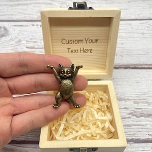 Op de afbeelding: Een bronzen kattenbeeldje met opgeheven armen wordt voor een open houten kistje gehouden. Op het kistje staat de tekst "Custom Your Text Here". De kist is gevuld met gescheurd papier.