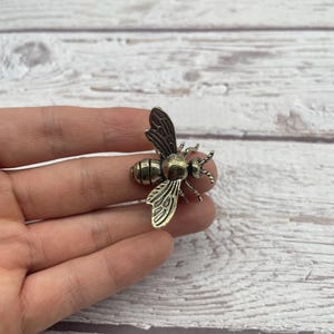 May include: A bronze-colored bee-shaped metal charm. The bee has detailed wings, body segments, and antennae. The charm is held in a hand against a white wood-grain background.