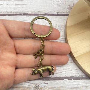 May include: Antique brass-toned keychain featuring a small pig charm and the letter "R". The keychain includes a split ring and a chain connecting the pig and letter charms. The pig charm is detailed with visible legs and snout.