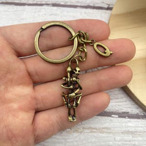 May include: Antique brass-colored skeleton keychain. The skeleton is holding a staff and is attached to a key ring and a small charm. The keychain is made of metal and has a vintage look.