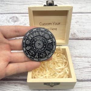 Può includere: Una ruota zodiacale in bianco e nero con simboli astrologici è tenuta davanti a una scatola di legno aperta. La scatola contiene carta sminuzzata e ha il testo "Custom Your Text Here" inciso.