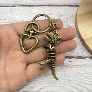 May include: An antique brass-toned keychain with a dinosaur skeleton charm, a heart-shaped clasp, and a circular keyring. The dinosaur charm has detailed rib and bone structures. An initial "H" charm hangs from the keyring.